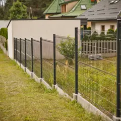 Nowoczesne ogrodzenie panelowe pod kątem wyznacza granicę posesji, za nim widać domy i zieleń.