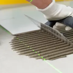 Ręka w rękawiczce rozprowadza klej pod płytki za pomocą kielni. Widać zielone linie lasera, które pomagają w precyzyjnym ułożeniu.