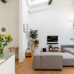 Jasny salon z szarymi sofami, telewizorem i roślinami. Wspaniały, wysoki sufit nad salonem dodaje przestrzeni.