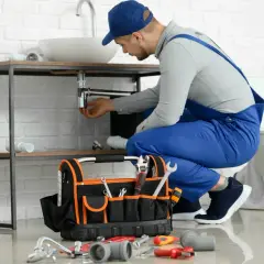 Hydraulik dokonuje naprawy spłuczki podtynkowej, pracując pod umywalką. Obok leży torba z narzędziami.