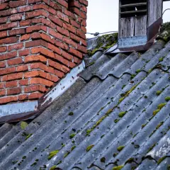 Stary dach sąsiada pokryty eternitem, z widocznym, ceglanym kominem i porostami. Azbest na dachu sąsiada budzi niepokój.