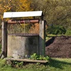 Stary, betonowy krąg studni kręgowej z drewnianą konstrukcją i metalowym kołowrotem. W tle jesienne drzewa i kopiec ziemi.