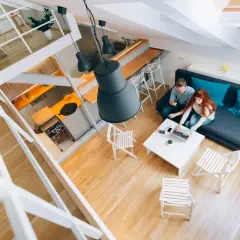 Para spędza czas w nowoczesnym salonie z otwartym sufitem. Kobieta pracuje na laptopie, mężczyzna obok niej pije wino.