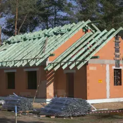 Budowa domu systemem gospodarczym. Dom z cegły, z zieloną więźbą dachową, w otoczeniu drzew. Na placu budowy materiały budowlane.