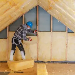 Pracownik w kasku montuje wełnę mineralną, wykonując ocieplenie poddasza nieużytkowego.