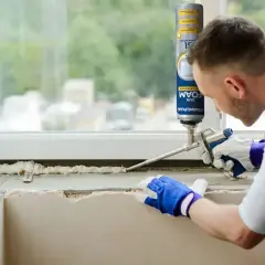 Mężczyzna w rękawiczkach nakłada piankę montażową na parapet. Czas, ile schnie pianka montażowa, zależy od warunków.