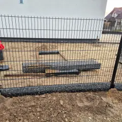 Elementy ogrodzenia panelowego w trakcie montażu. Widać słupki, panele i materiały budowlane na placu budowy.