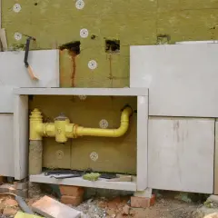 Widoczna przebudowa instalacji gazowej bez pozwolenia. Żółte rury i zawory w izolacji, obok kamienne płyty elewacyjne.