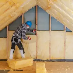 Pracownik w kasku i masce izoluje poddasze wełną mineralną. To etap prac nad niezgłoszona adaptacja poddasza, która ma zapewnić komfort cieplny.