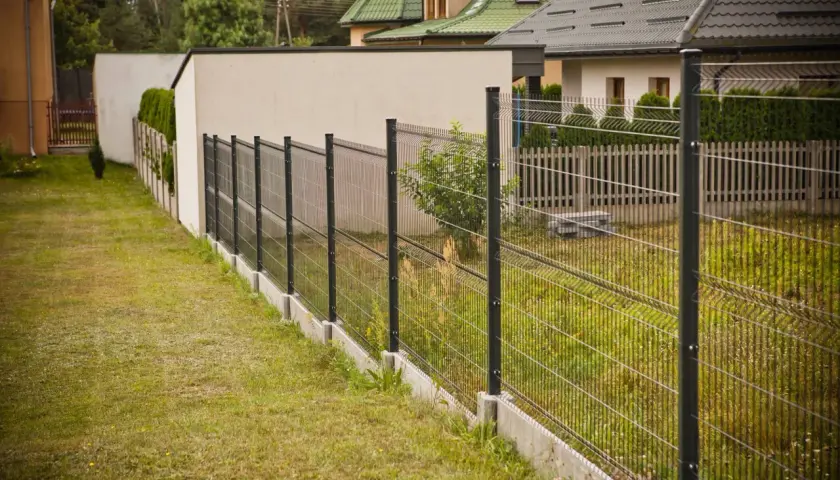 Nowoczesne ogrodzenie panelowe pod kątem wyznacza granicę posesji, za nim widać domy i zieleń.