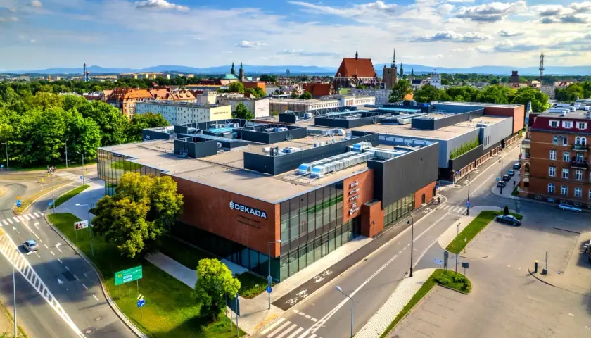 Nowoczesny budynek usługowy "Dekada" z przeszkloną fasadą i ceglanymi elementami, otoczony zielenią i drogami.