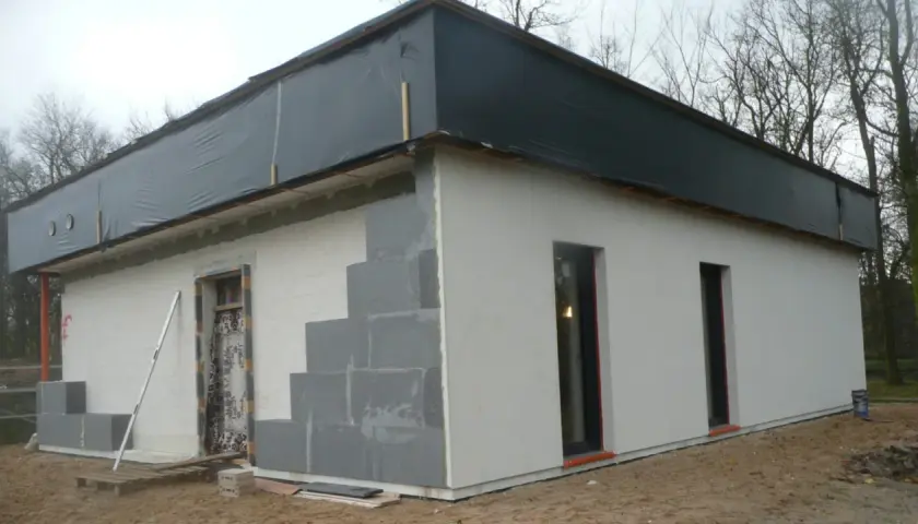 Budowa domu ze styropianu w toku. Ściany z bloczków, częściowo otynkowane, dach przykryty czarną folią.