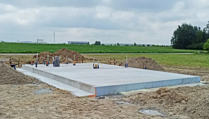 Gotowy fundament pod dom szkieletowy, z wystającymi rurami i zbrojeniem, otoczony piaskiem i zielonym polem.