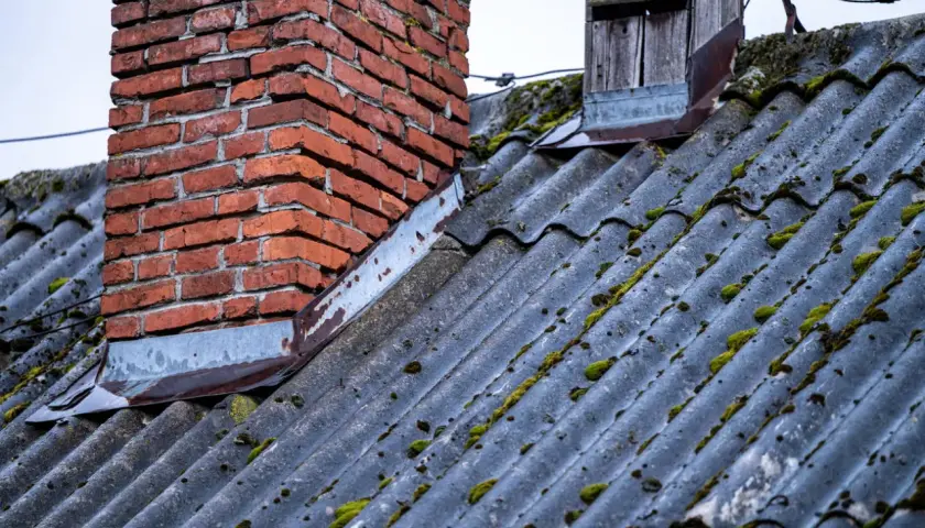 Stary dach sąsiada pokryty eternitem, z widocznym, ceglanym kominem i porostami. Azbest na dachu sąsiada budzi niepokój.