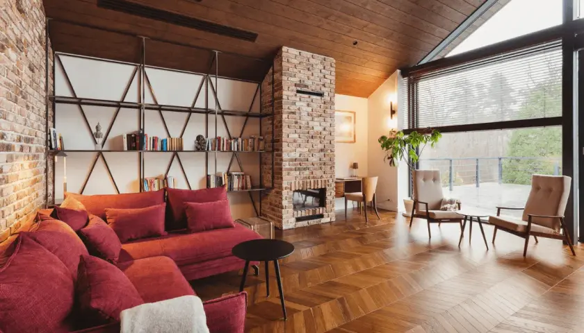 Przytulny salon w stylu wiejskim z ceglanym kominkiem, czerwoną kanapą i drewnianą podłogą.