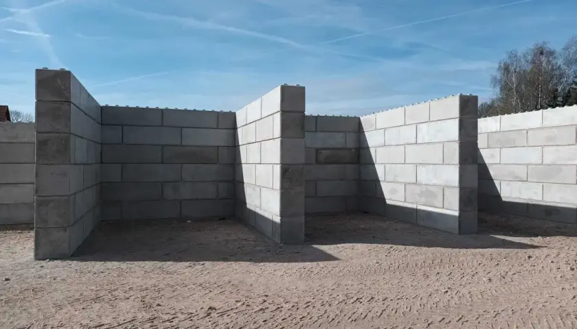 Budowa muru oporowego z bloczków betonowych. Konstrukcja tworzy zarys pomieszczeń na piaszczystym placu budowy pod błękitnym niebem.