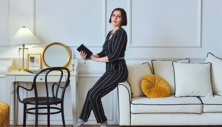 Kobieta w eleganckim, prążkowanym stroju siedzi na kanapie, trzymając książkę. To kwintesencja stylu glamour – wyrafinowanie i blask.