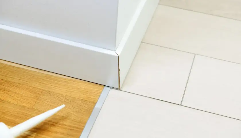 Łączenie płytek z panelami korkiem. Widać białą listwę przypodłogową, drewnianą podłogę i jasne panele.
