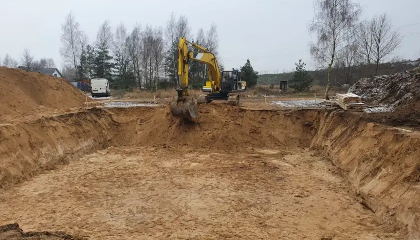 Koparka pracuje przy wykopie pod fundament. Piasek i ziemia tworzą głęboki dół, przygotowany do dalszych prac budowlanych.
