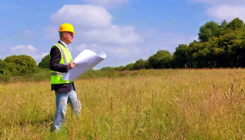 Inżynier w kasku i kamizelce odblaskowej analizuje plany na tle łąki, przygotowując się do badań geotechnicznych gruntu.