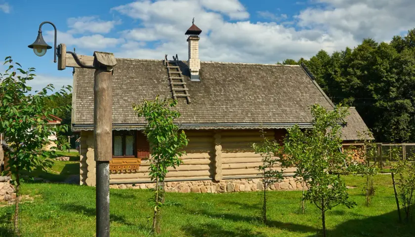 Drewniany dom na działce rolnej, z kamiennym fundamentem i gontowym dachem. Wokół młode drzewka i trawa.