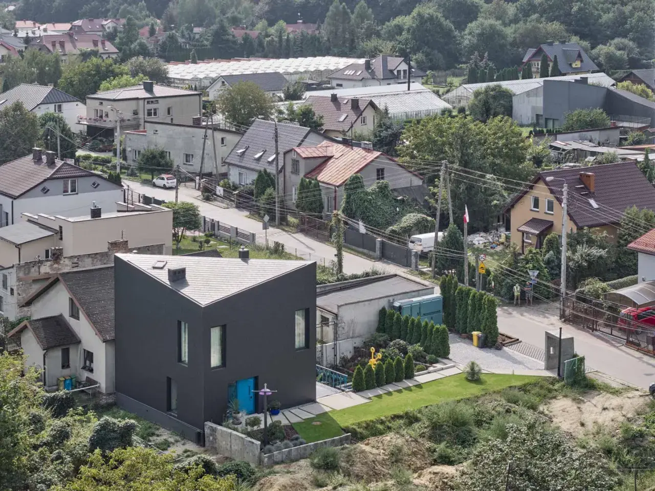 Nowoczesny dom o trójkątnym kształcie na tle zabudowy jednorodzinnej. Widać tu typowe **grunty rolne zabudowane**, z domami, szklarniami i zielenią.