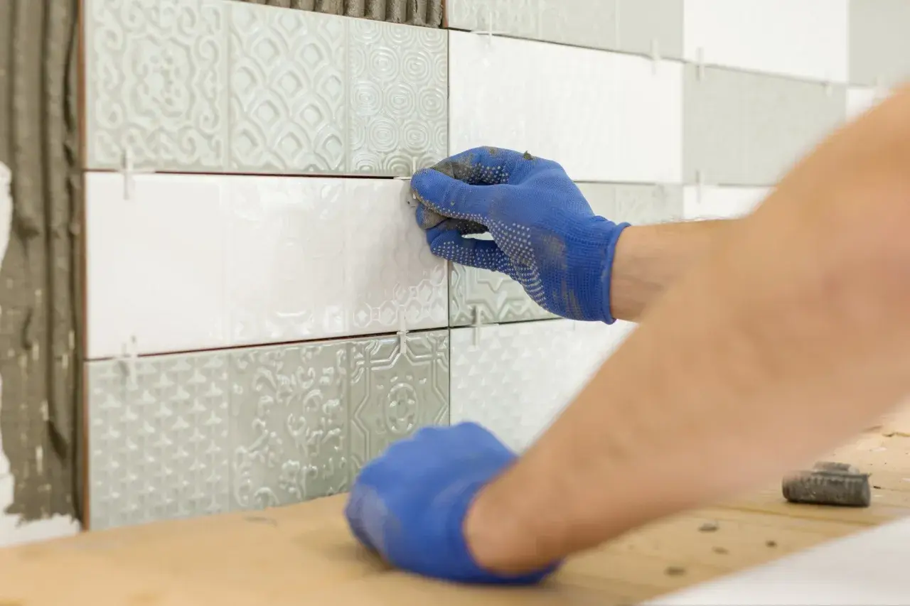 Ręce w niebieskich rękawiczkach wykonują naprawę płytek ceramicznych, precyzyjnie układając nowe elementy na ścianie.