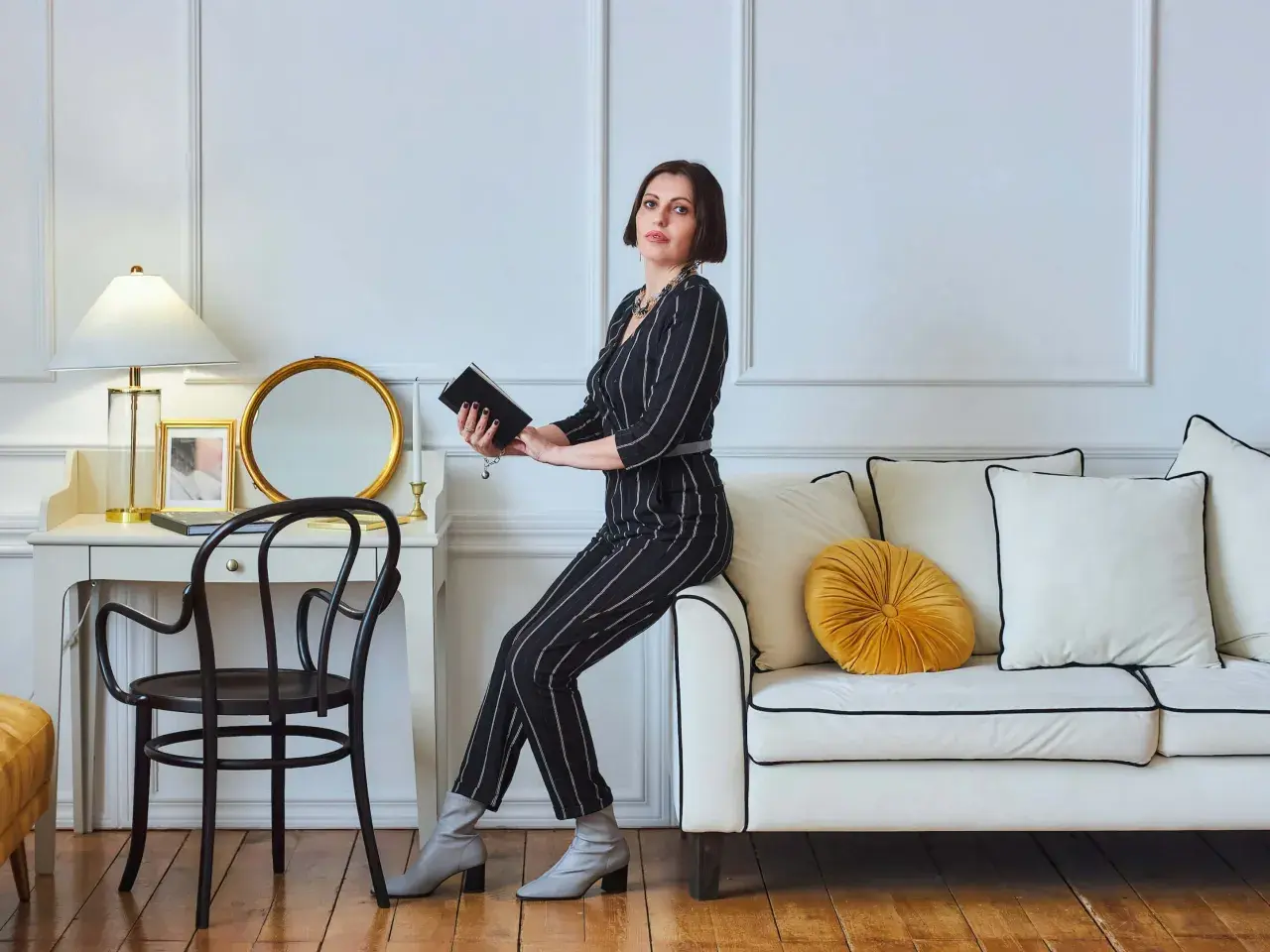 Kobieta w eleganckim, prążkowanym stroju siedzi na kanapie, trzymając książkę. To kwintesencja stylu glamour – wyrafinowanie i blask.