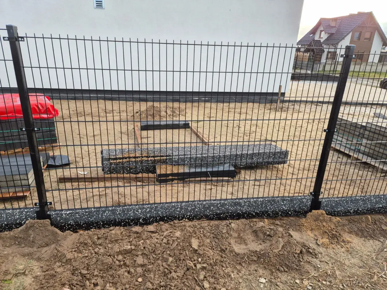 Elementy ogrodzenia panelowego w trakcie montażu. Widać słupki, panele i materiały budowlane na placu budowy.