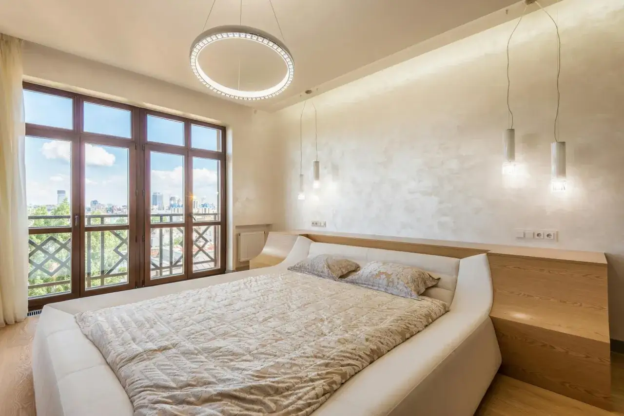 Przytulna sypialnia inspiracje: nowoczesne łóżko, okna z widokiem na miasto, okrągła lampa i subtelne oświetlenie tworzą relaksującą atmosferę.