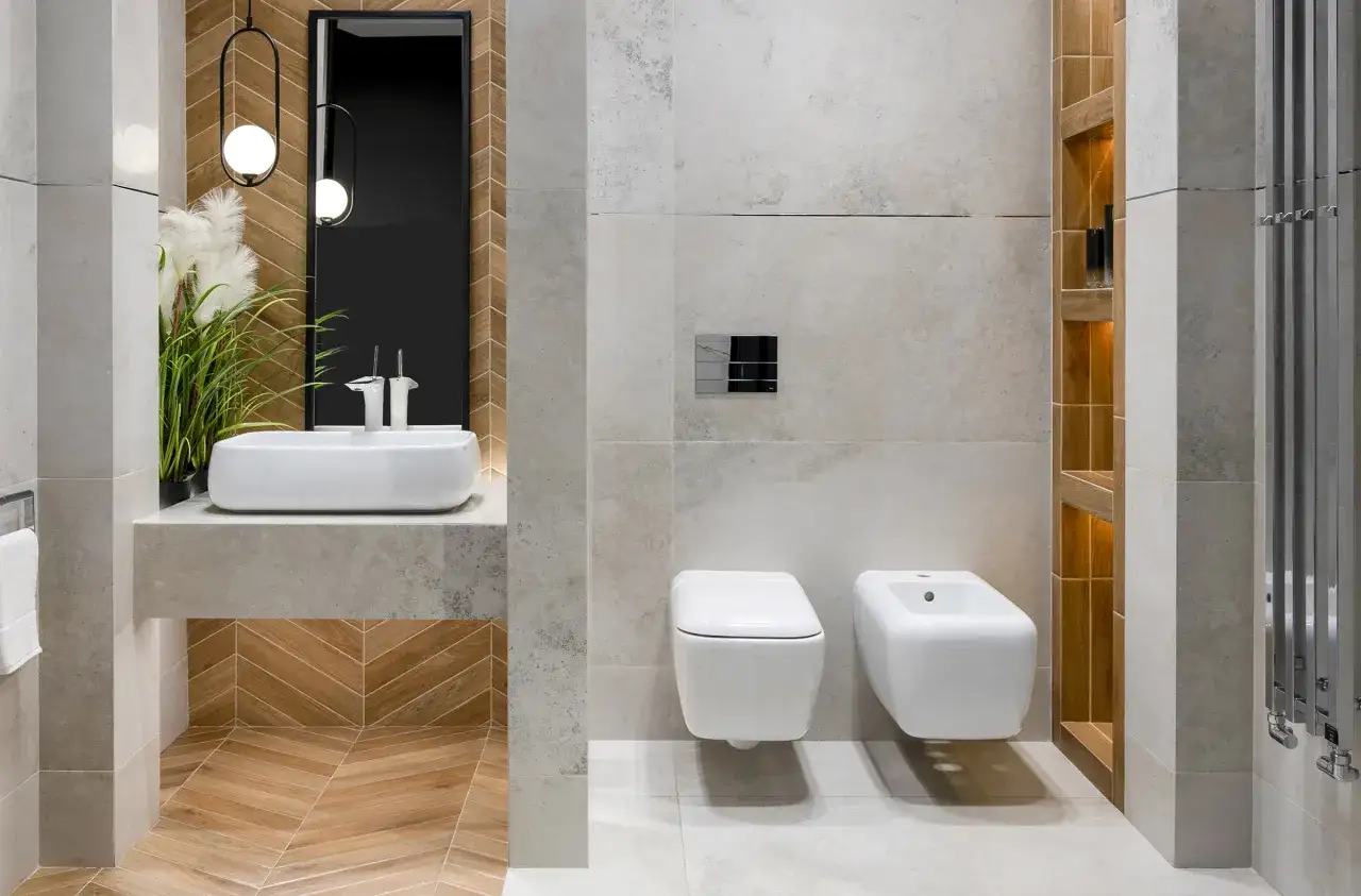 Nowoczesne układanie płytek w łazience: wisząca miska WC i bidet na tle kamiennych płytek, umywalka nablatowa i drewniana podłoga w jodełkę.