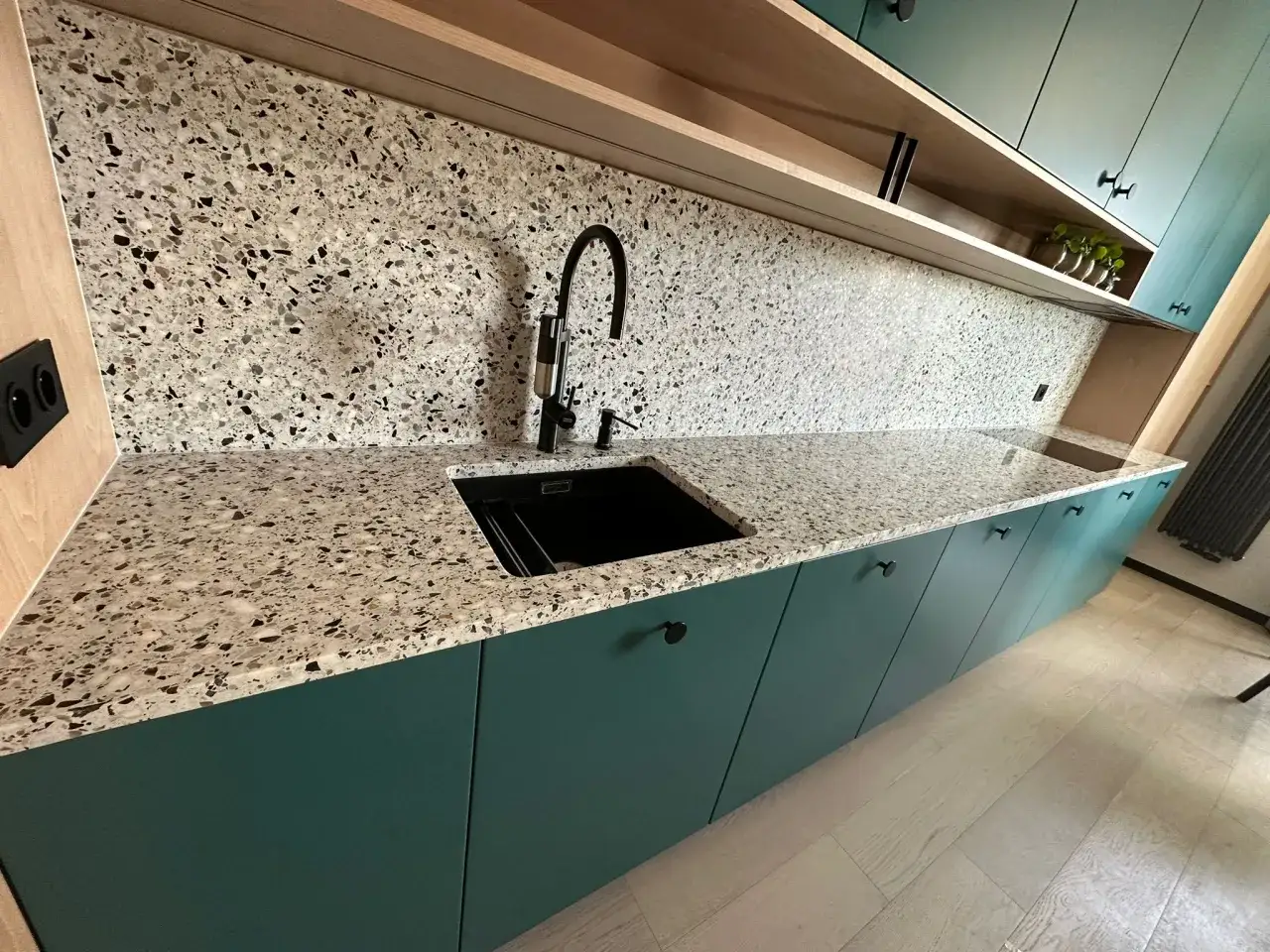 Nowoczesna kuchnia z zielonymi szafkami i blatem z lastryko. Czarny zlew i bateria dodają elegancji.