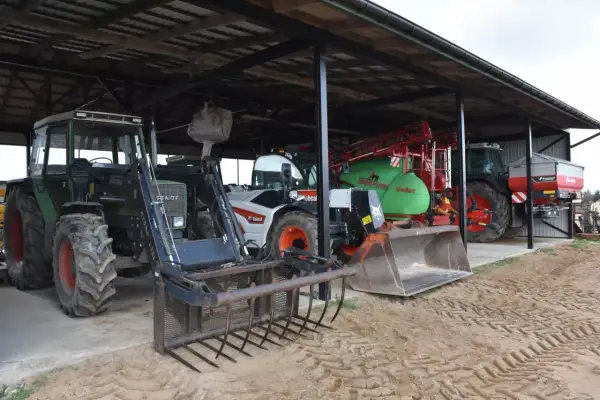 Ciągniki i maszyny rolnicze pod zadaszeniem. Widać zielony Fendt z ładowaczem, biały Bobcat, zielony opryskiwacz i ciągnik z rozsiewaczem. To wszystko w budynku gospodarczym bez zgłoszenia.